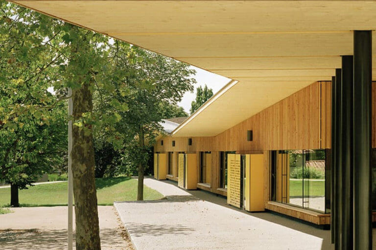 Architecture durable et éco-construction : école en bois-paille. Il y a trois arbres sur la gauche et l'école sur la droite. agence Tectonique Architectes Lyon